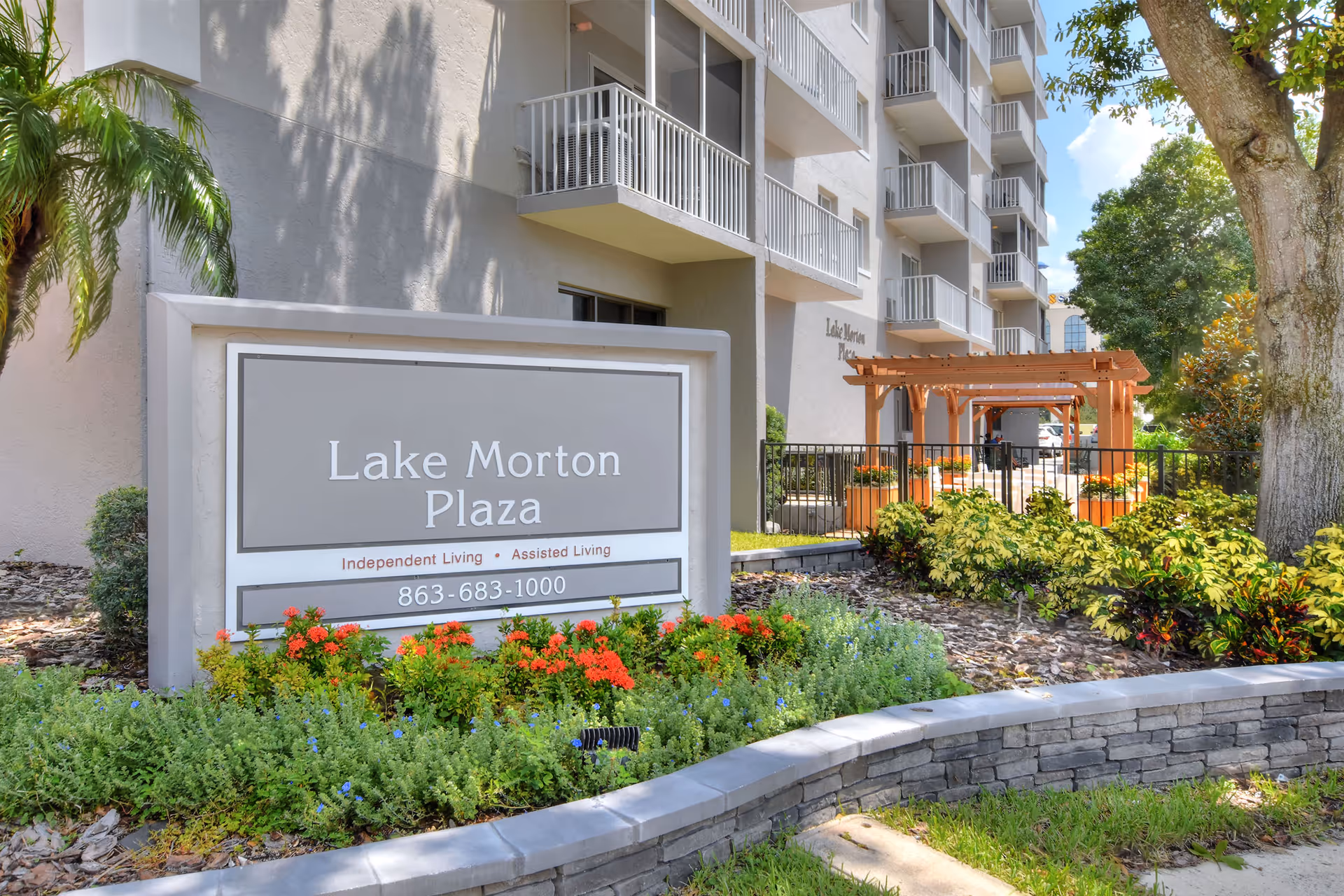 Outdoor view of Lake Morton Plaza senior living facility sign surrounded by colorful flowers and greenery, with the multi-story building and balconies in the background under a sunny sky.
