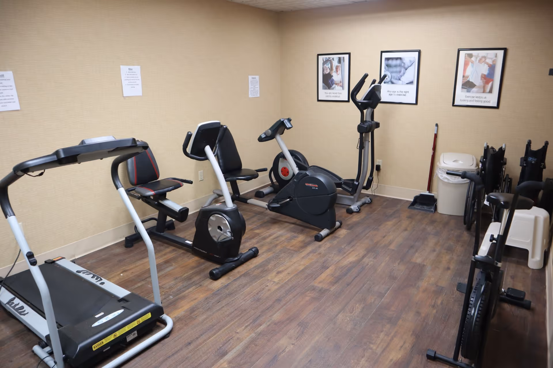 A small fitness room with exercise equipment including a treadmill, two stationary bikes, and an elliptical machine. The room has wood flooring, beige walls, and three framed motivational posters on the wall. There is a trash can and some folded chairs in the corner.