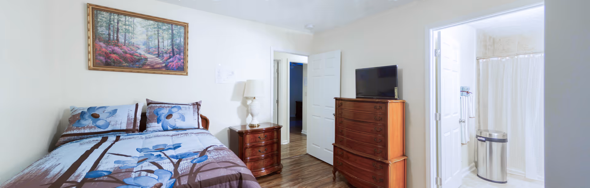 A bedroom with a bed covered in blue floral bedding, a wooden nightstand with a lamp, a wooden dresser with a flat-screen TV on top, and an open door leading to a bathroom with a shower curtain and a stainless steel trash can.