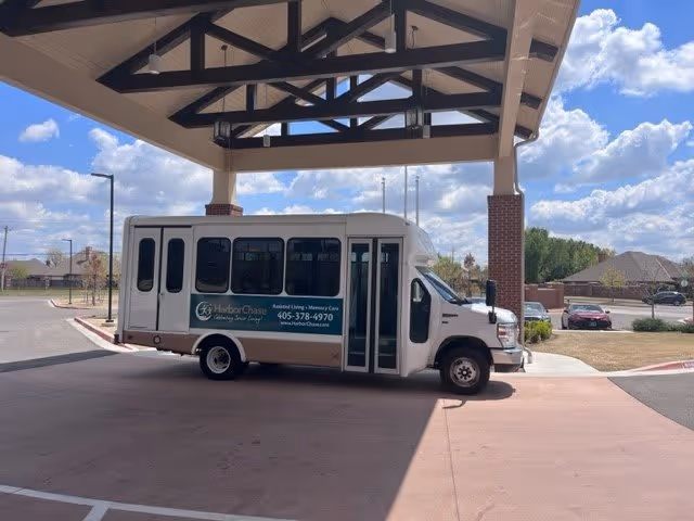 A white and beige shuttle bus parked under a covered drop-off area at a senior living facility. The bus has signage for HarborChase Assisted Living and Memory Care with a phone number and website. The background shows a partly cloudy sky, street lamps, and residential houses.
