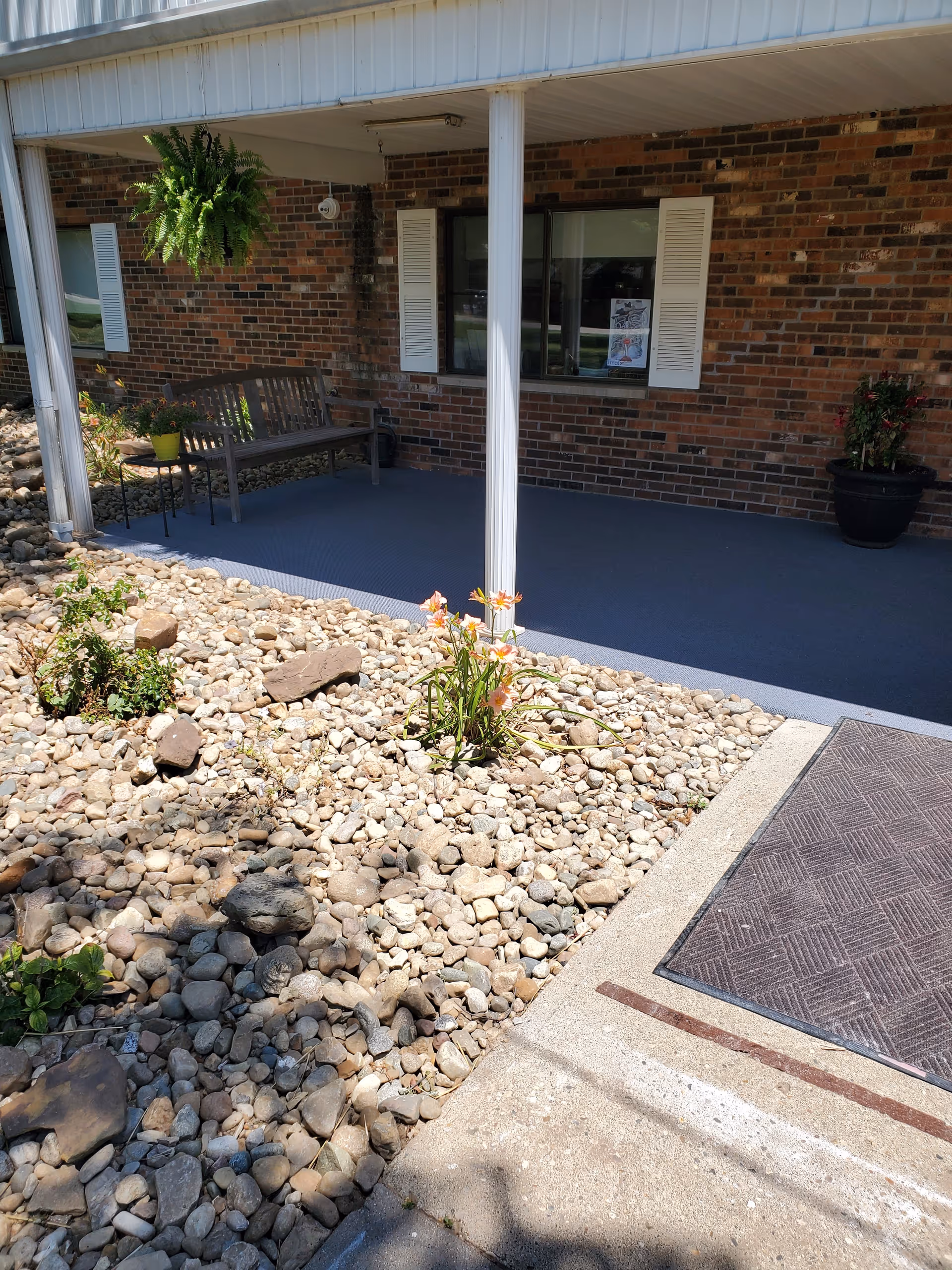 Covered front porch of a brick building with white columns, a bench, potted plants, and rock landscaping.