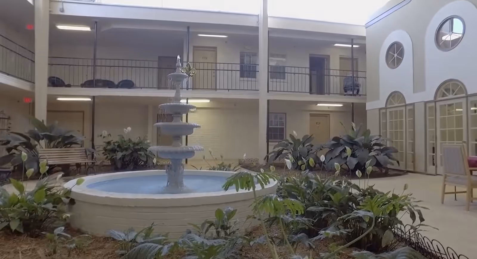 Indoor courtyard area with a multi-tiered water fountain in the center, surrounded by green plants and benches. The courtyard is enclosed by two levels of rooms with doors and windows, and there are large windows and doors on one side letting in natural light.