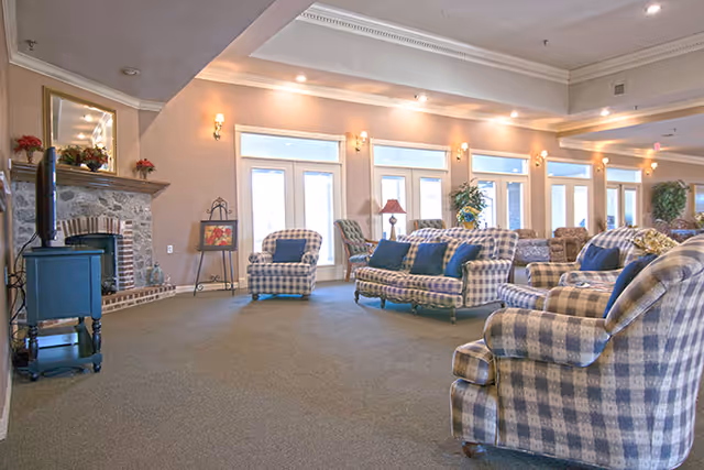 A spacious living room area with multiple plaid upholstered armchairs and sofas arranged around a stone fireplace with a TV on a stand. The room has large windows and glass doors letting in natural light, beige walls with sconces, and a carpeted floor.