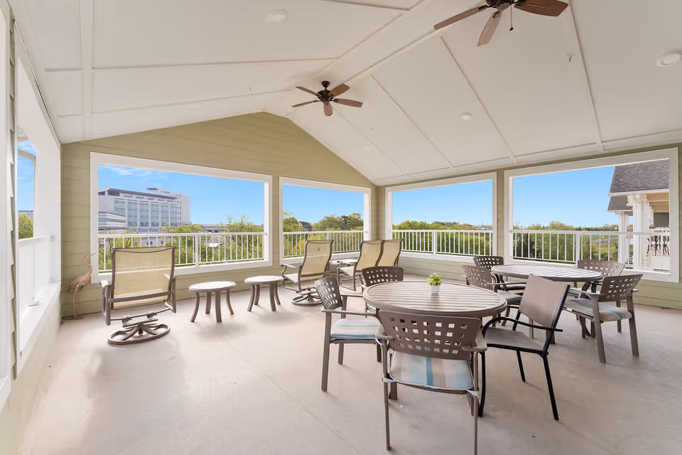 Covered outdoor patio area with several round tables and chairs, some cushioned rocking chairs, and large open windows offering a view of greenery and a distant building under a clear blue sky.