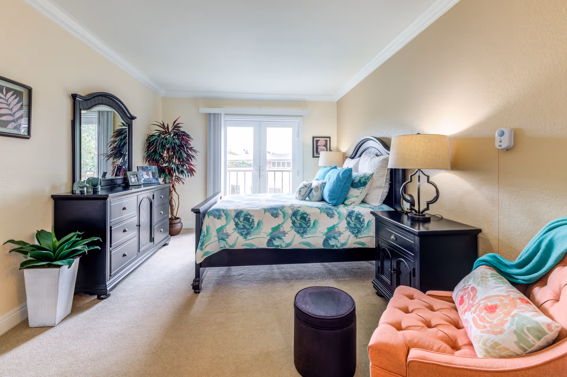 A bright and cozy bedroom with a large bed featuring floral bedding and multiple pillows. The room includes a black dresser with a mirror, a bedside table with a lamp, a coral-colored armchair with a floral pillow and teal throw, a small round ottoman, and two potted plants. French doors with vertical blinds lead to a balcony or outside area, allowing natural light to fill the room.