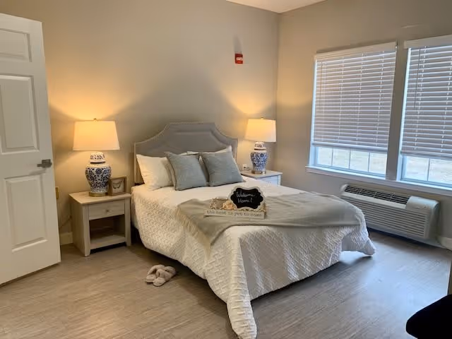 A cozy bedroom in a senior living facility with a neatly made bed featuring white bedding and multiple pillows. There are two bedside tables, each with a blue and white lamp. A small decorative sign on the bed reads 'Welcome Home! the best is yet to come.' The room has light-colored walls, a window with blinds, and a heating/cooling unit beneath the window. A pair of slippers is placed on the floor near the bed.