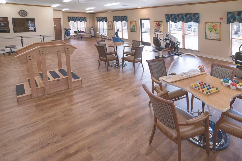 Bright multipurpose activity and therapy room with wood floors, tables and chairs, exercise equipment, and therapy stairs.