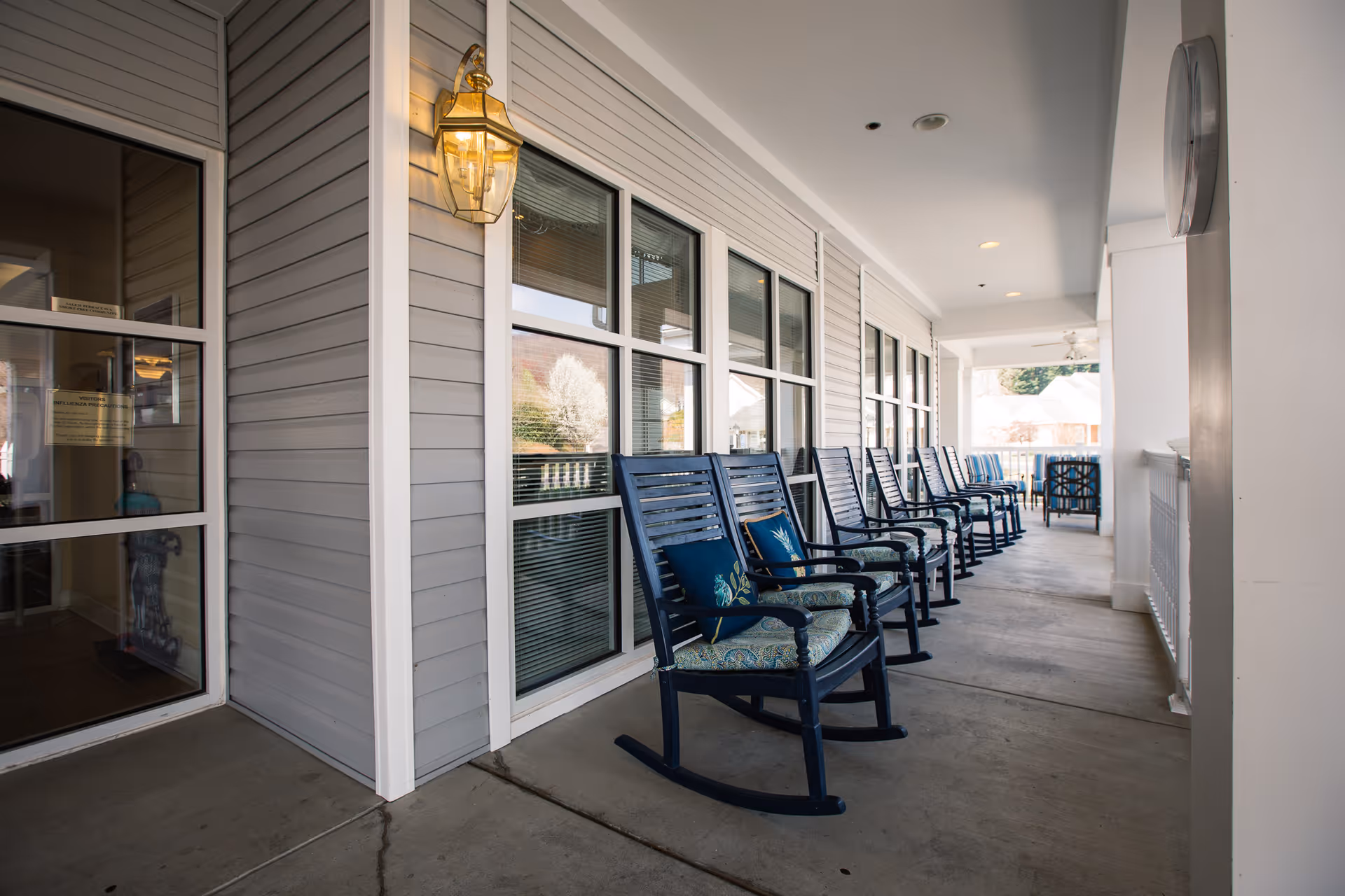 A covered outdoor porch area at Friendship Salem Terrace with a row of blue rocking chairs, each with cushions and pillows, lined up against the wall with windows. A lantern-style light fixture is mounted on the wall near the entrance door.