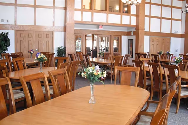 A spacious dining room with multiple wooden tables and chairs arranged neatly. Each table has a small vase with colorful flowers. The room features wood paneling on the walls and large windows allowing natural light to enter.