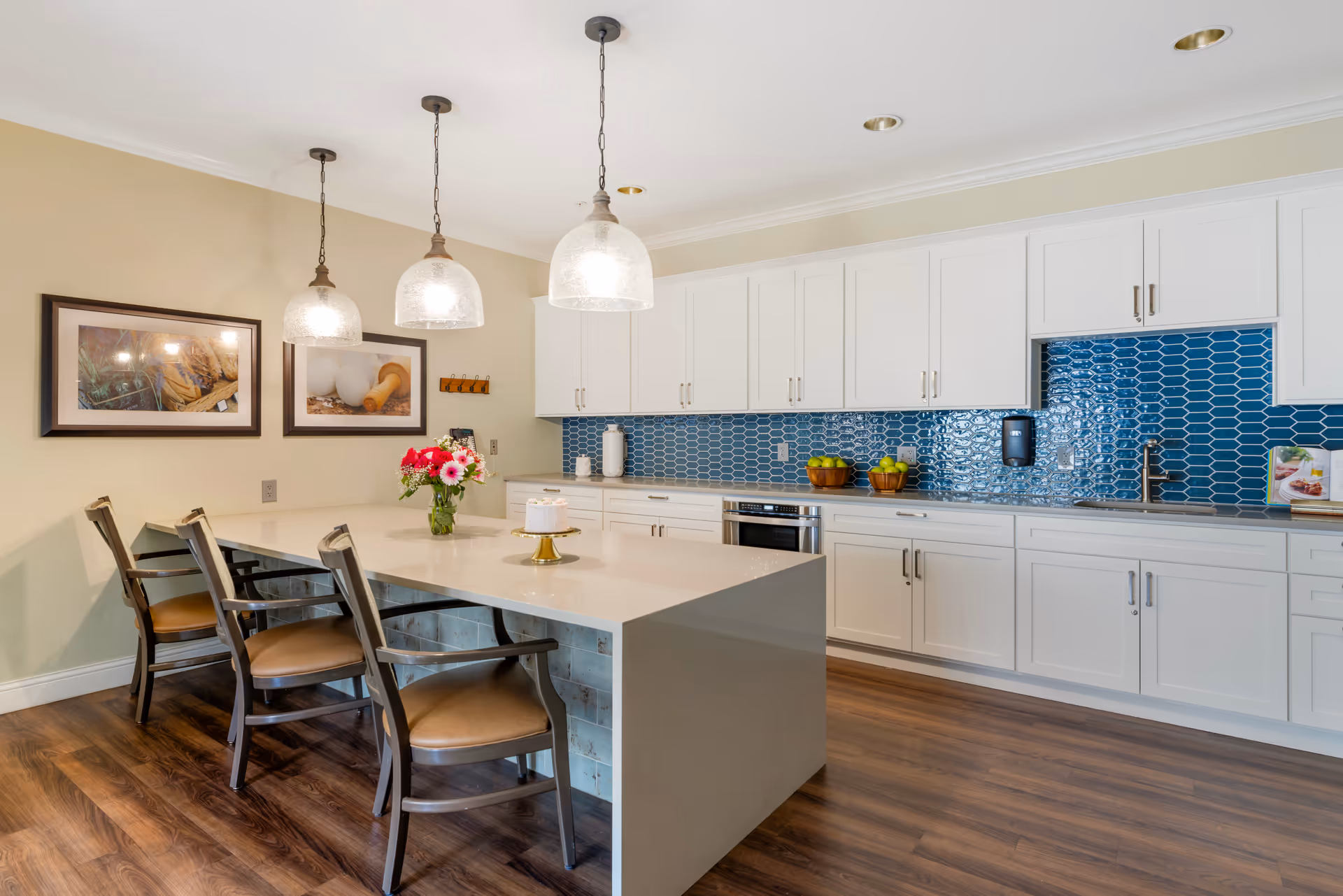 A modern kitchen area with a long white island countertop and four wooden chairs with armrests. Three pendant lights hang above the island. The back wall features white cabinets and a blue tiled backsplash. There are decorative items including a vase with flowers, a cake on a stand, and bowls of fruit on the countertop. The floor is wooden, and two framed pictures hang on the beige wall to the left.