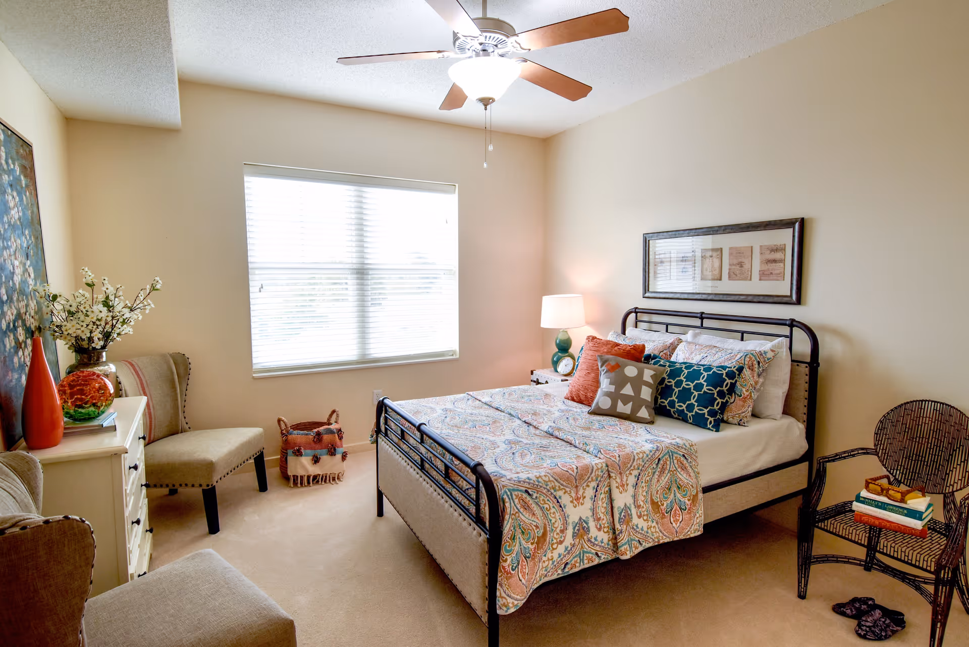 A bright bedroom with a large window covered by blinds, a bed with a patterned bedspread and multiple decorative pillows, a ceiling fan with a light, a nightstand with a lamp, a wicker chair with books on it, two beige upholstered chairs, a small white dresser with decorative items including a vase with flowers and a red vase, and a framed picture on the wall above the bed.