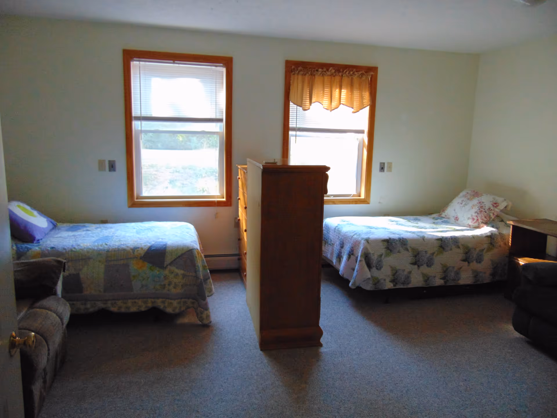 A bedroom with two single beds separated by a tall wooden dresser. Each bed has a patterned quilt and a pillow. There are two windows with wooden frames behind the beds, one with blinds and the other with a valance. The room has carpeted flooring and neutral-colored walls.