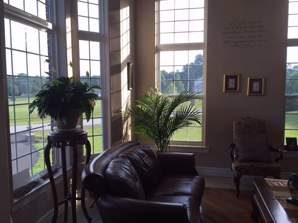 A cozy living room area with large windows letting in natural light. There is a dark leather couch, a floral upholstered armchair with a cushion, a wooden side table with a potted plant, and another large green plant near the window. The walls have framed pictures and a wall decal with an inspirational quote. Outside the windows, a green lawn and trees are visible.