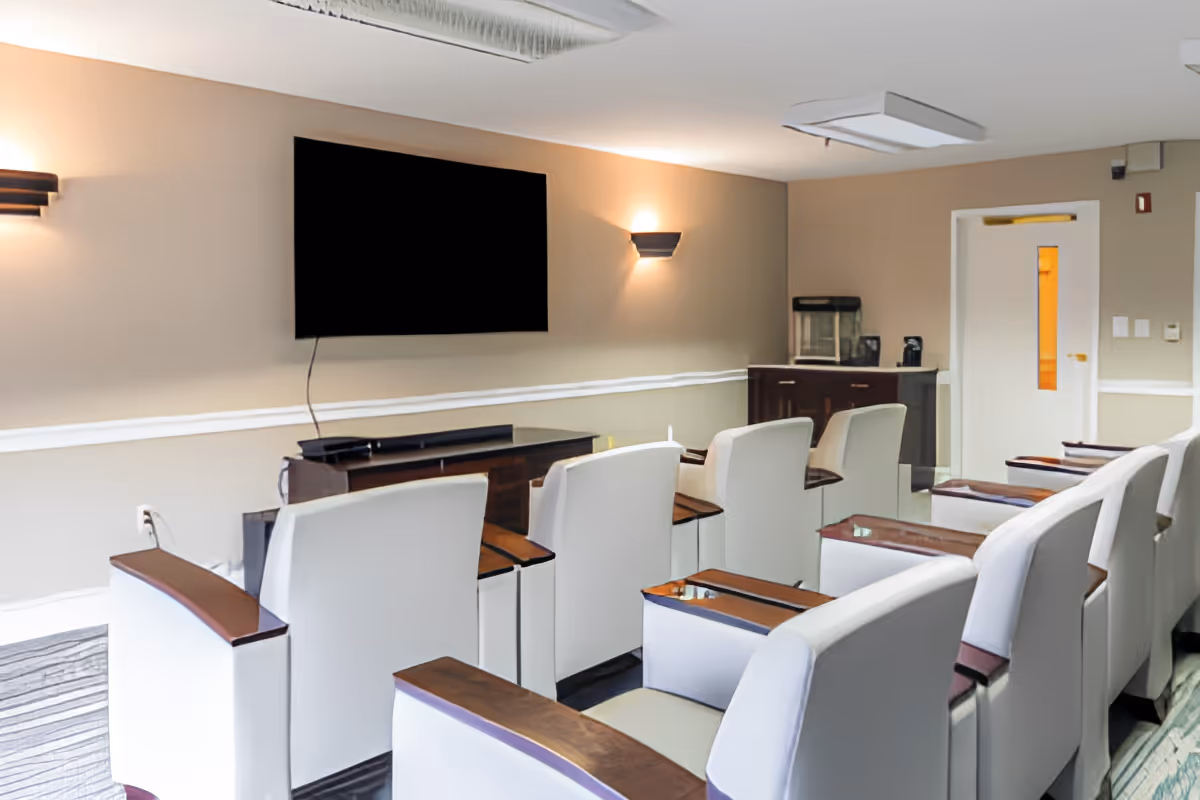 A small theater-style room with two rows of white armchairs featuring wooden armrests, all facing a large flat-screen TV mounted on a beige wall. The room has soft wall sconces for lighting, a cabinet with a popcorn machine and coffee maker in the corner, and a door with a narrow window on the right side.