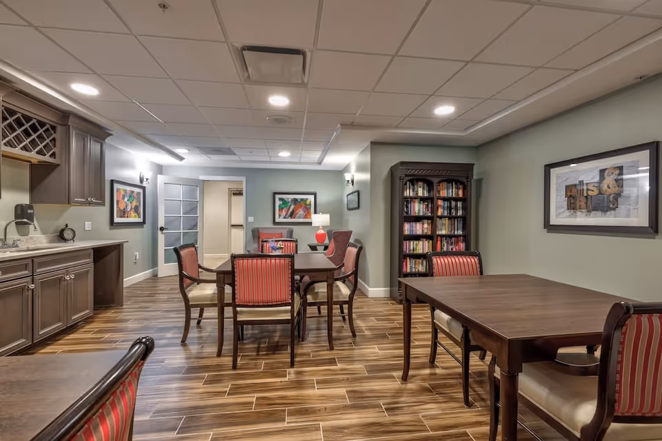 Interior view of a common area in a senior living facility featuring wooden tables and chairs with red and beige striped cushions, a bookshelf filled with books, framed artwork on the walls, wood-look tile flooring, and a kitchenette area with cabinets and a countertop on the left side.