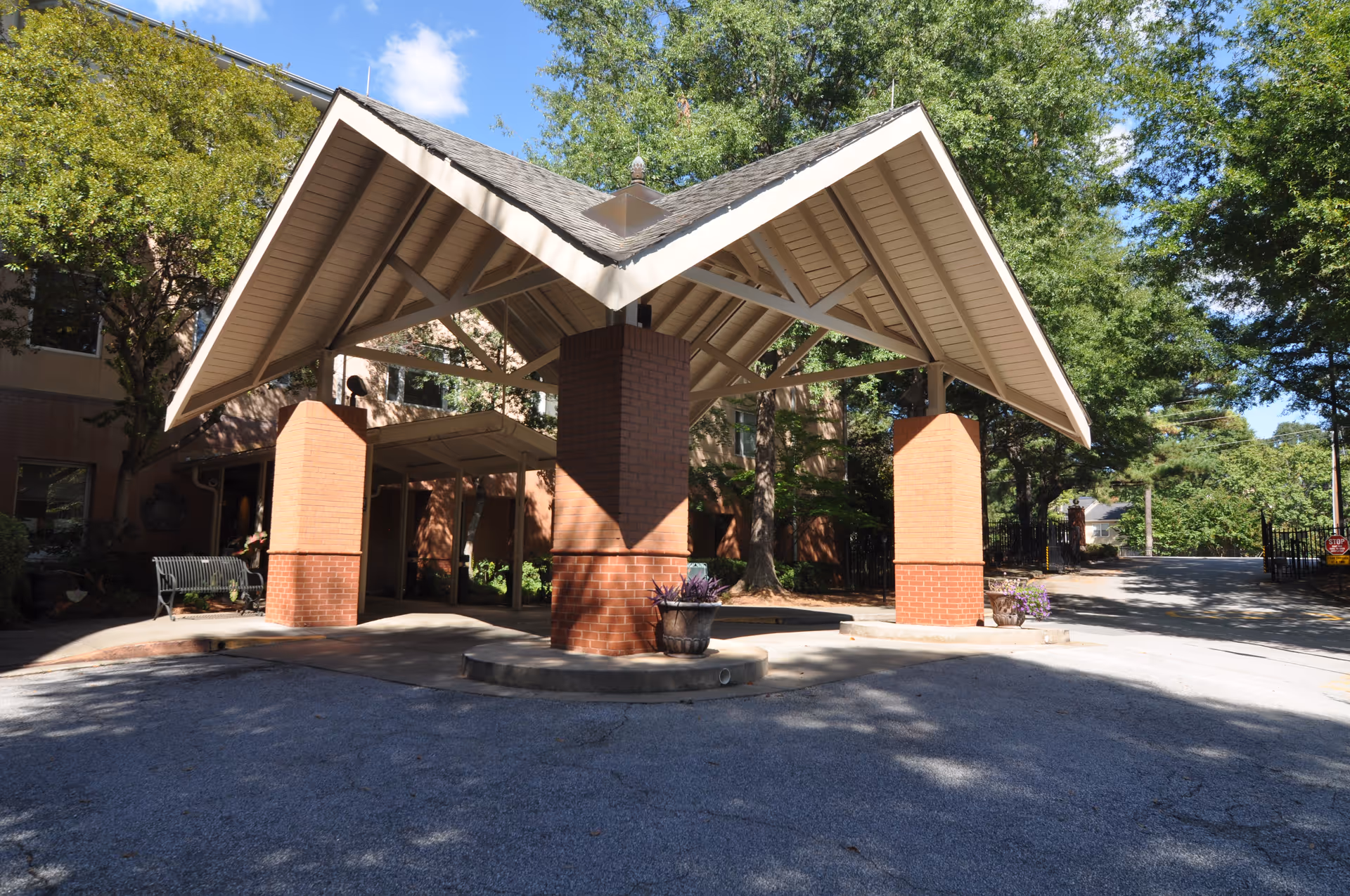 Covered entrance area with a peaked roof supported by four brick pillars at a senior living facility. The surrounding area includes trees, a bench, and a driveway leading to the entrance.