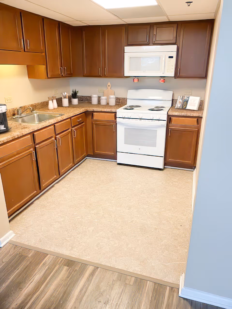 A kitchen with brown wooden cabinets, a white stove with an oven, and a white microwave mounted above the stove. The countertop is a speckled brown color with various kitchen items including a coffee maker, small plants, and containers. The floor transitions from wood to a beige linoleum surface.