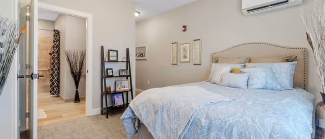 A cozy bedroom with a neatly made bed featuring light blue patterned bedding. The room has beige walls with three framed pictures hanging above the bed. To the left, there is an open doorway leading to a hallway with light wood flooring and a tall vase with decorative branches. A black ladder-style shelf holds framed photos and decor items. An air conditioning unit is mounted on the wall above the bed.
