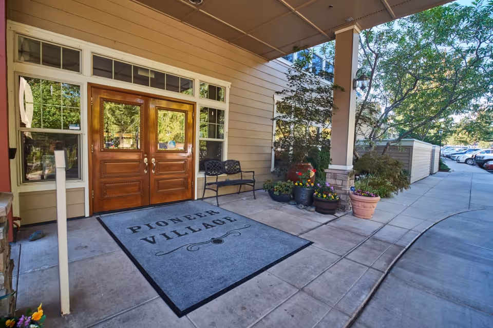 Covered entrance to Pioneer Village with double wooden doors, a welcome mat reading 'Pioneer Village', a bench, and potted plants.