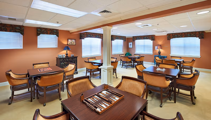 A well-lit common room with multiple tables and chairs arranged for group activities or dining. The walls are painted a warm orange color, and windows with blinds and floral valances let in natural light. There are lamps on small cabinets along the walls and a backgammon game set up on the table in the foreground.