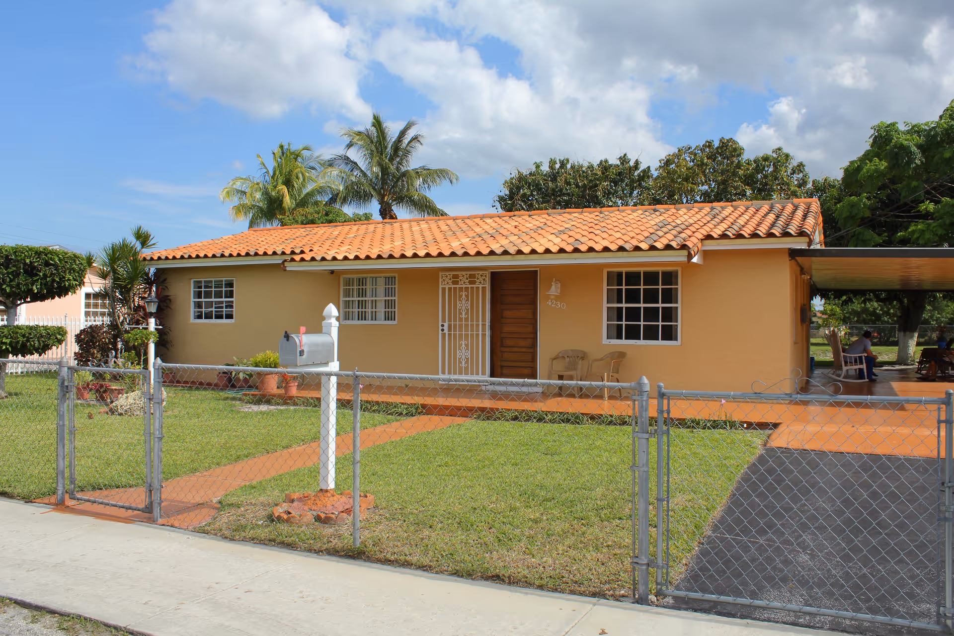 Single-story house with a terracotta tiled roof, beige exterior walls, and a fenced front yard with a mailbox and a concrete pathway leading to the front door. There are two chairs on the front porch and palm trees in the background under a partly cloudy sky.