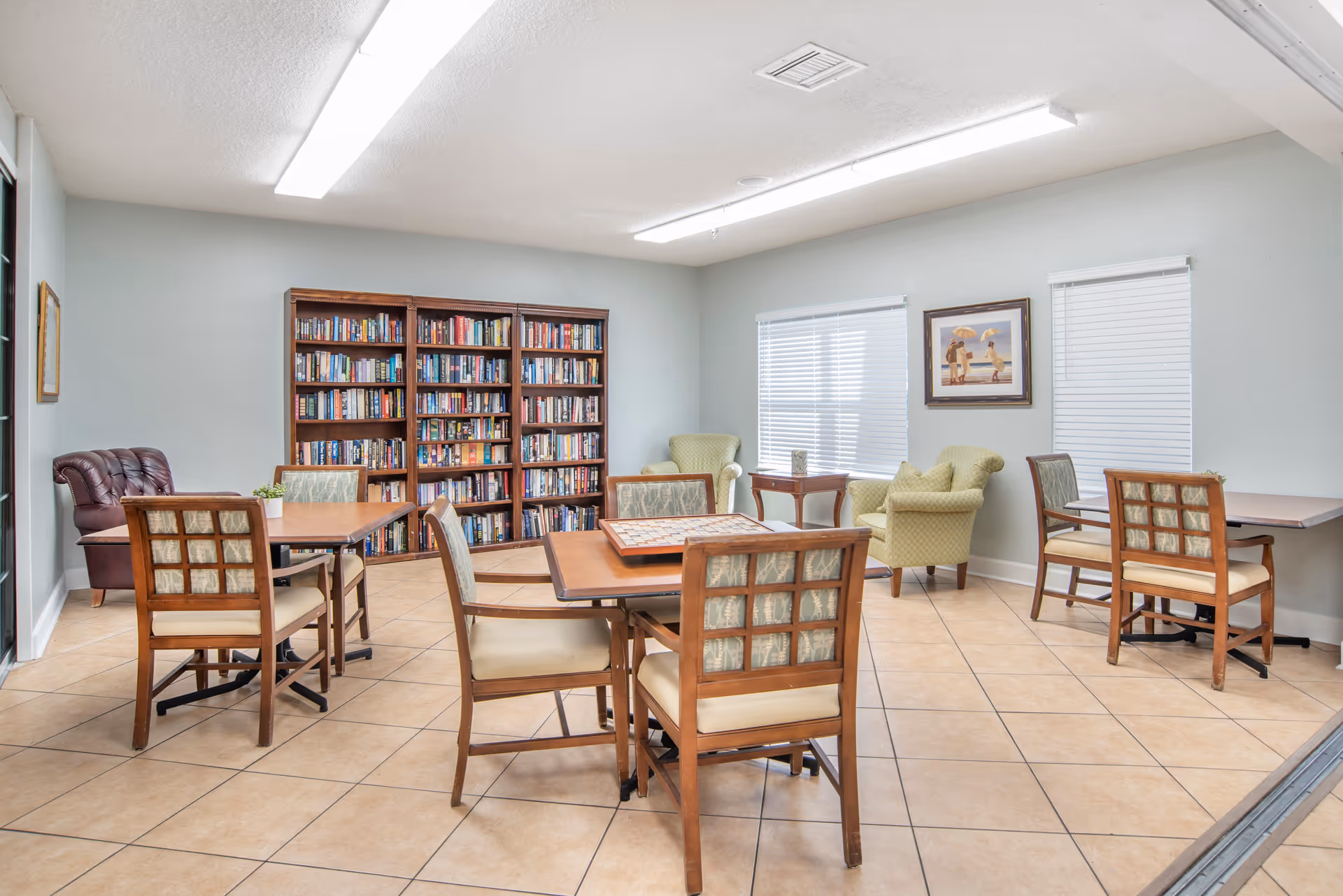 A bright room with tiled floors featuring several wooden tables and chairs arranged for seating. There is a large bookshelf filled with books against one wall, two green upholstered armchairs near two windows with blinds, and a framed picture on the wall. The room is well-lit with fluorescent ceiling lights.