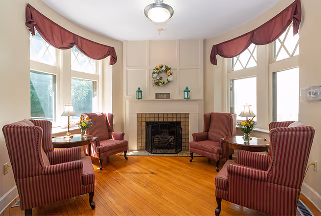 A cozy living room with four red and white striped armchairs arranged around two small round wooden tables with flower vases. The room features a fireplace with a decorative wreath and two lanterns on the mantel. Large windows with red valances allow natural light to fill the space, and the floor is wooden.