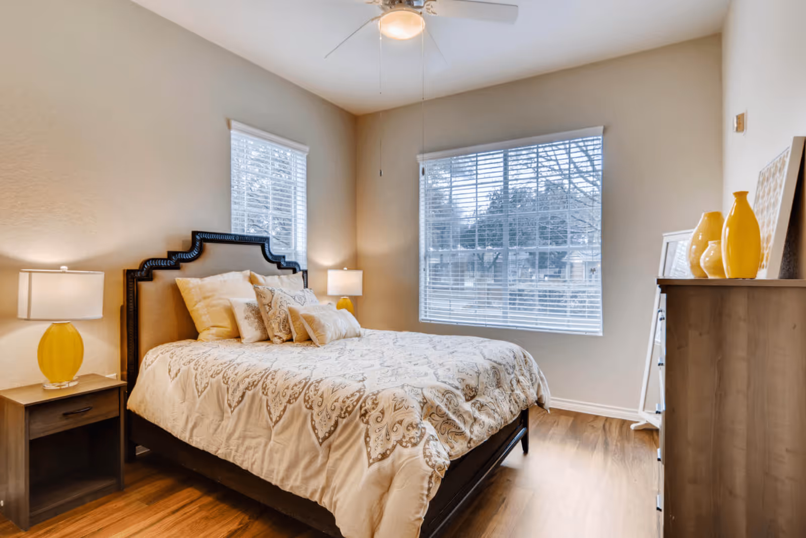 A cozy bedroom with a large bed featuring a decorative headboard and patterned bedding. There are two windows with white blinds letting in natural light. On either side of the bed are nightstands with yellow lamps. A wooden dresser with yellow vases and framed artwork is positioned against the wall, and a ceiling fan with a light is mounted above.