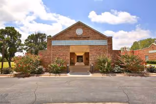 Front exterior view of a brick building with a peaked roof and an entrance walkway flanked by bushes and trees under a partly cloudy sky.