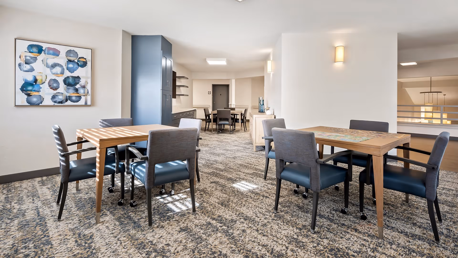 A bright and spacious common area with multiple wooden tables and gray cushioned chairs on wheels arranged on a patterned carpet. The room has white walls, modern wall sconces, and abstract artwork hanging on one wall. In the background, there is a cabinet and more seating, with an open railing overlooking a lower level.