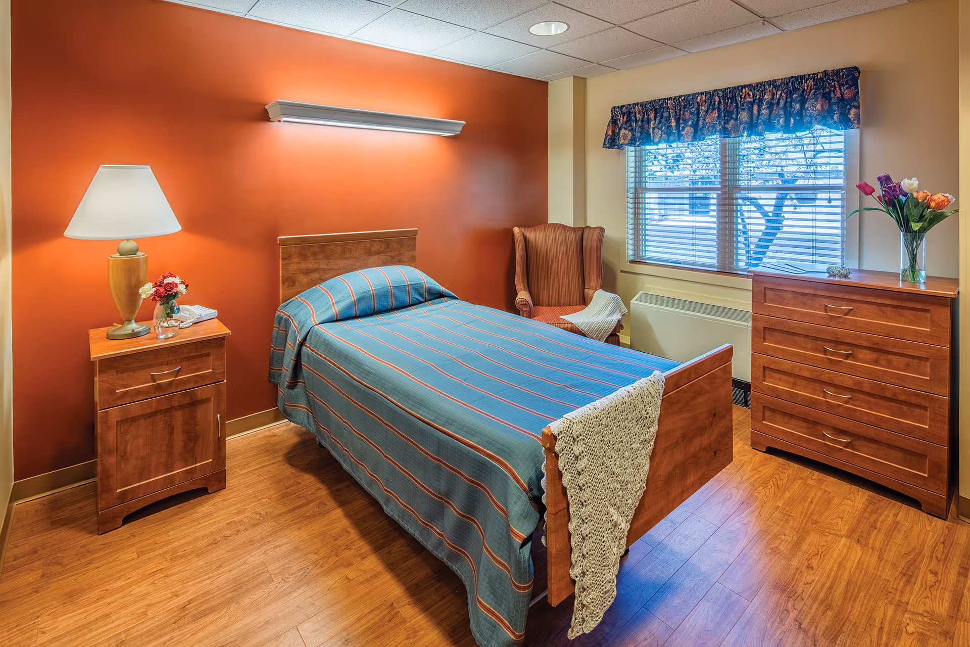 A cozy bedroom in a senior living facility with a single bed covered in a blue and orange striped bedspread. The room features wooden furniture including a nightstand with a lamp and flowers, a chest of drawers with a vase of colorful tulips, and a comfortable upholstered armchair near a window with blinds and a floral valance. The walls are painted in warm tones, with one accent wall in orange.