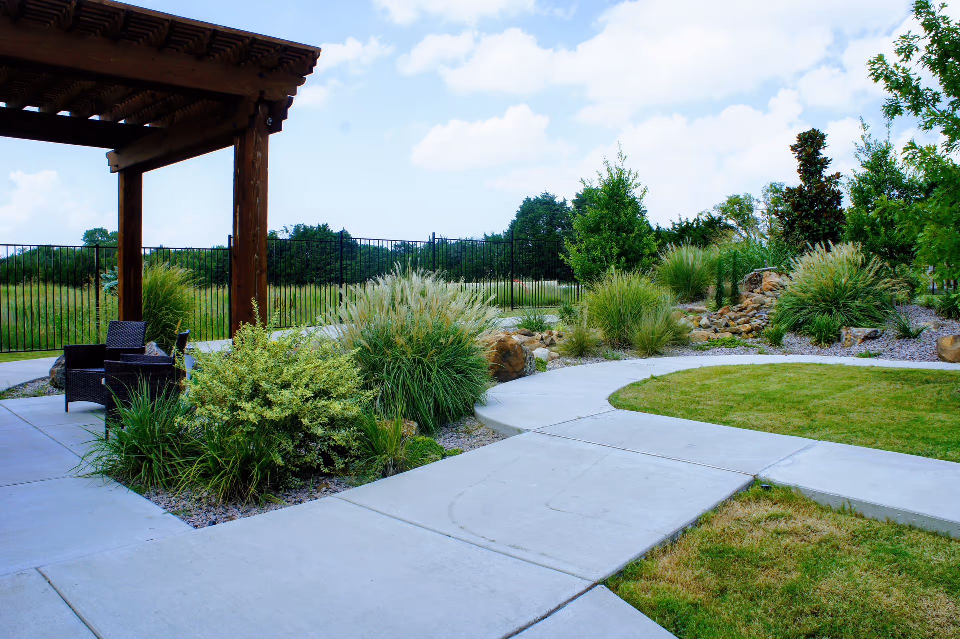 Outdoor garden area with a wooden pergola, patio chairs, various green shrubs and grasses, a curved concrete walkway, and a black metal fence in the background under a partly cloudy sky.