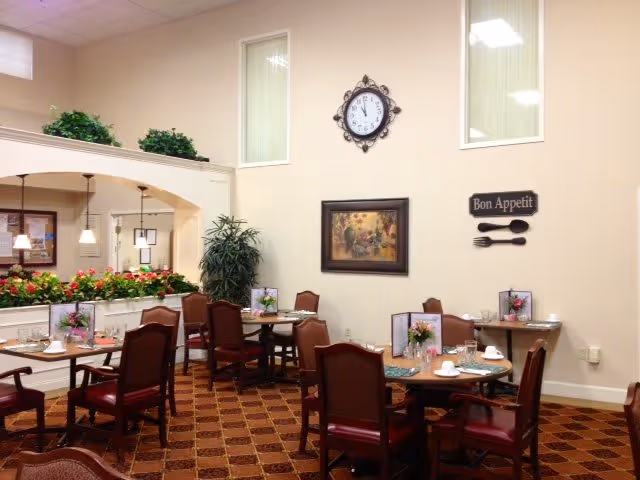 Assisted living dining room with multiple set tables and chairs, wall clock, artwork, plants, and a 'Bon Appetit' sign.