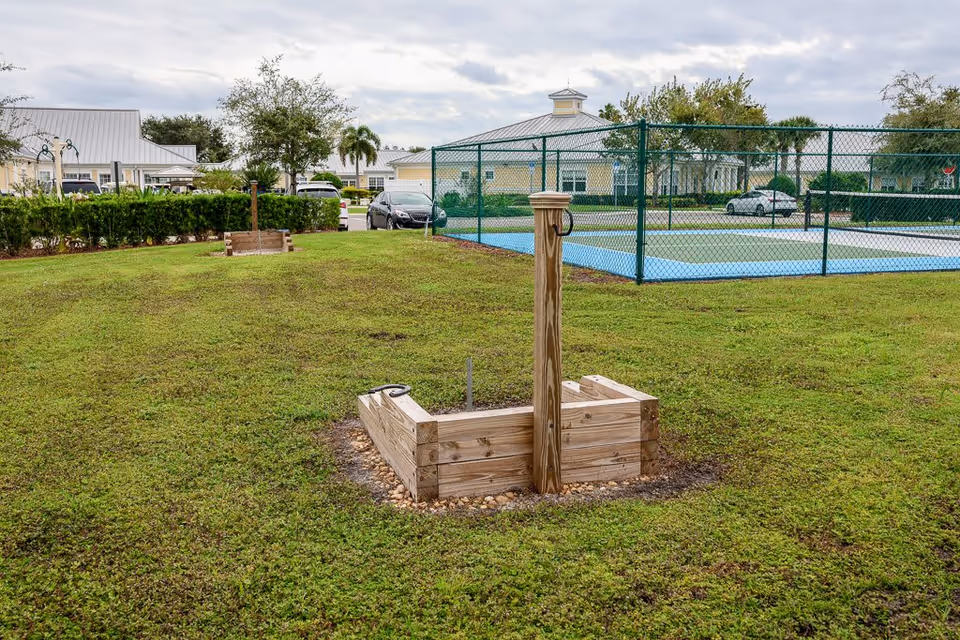 Outdoor grassy area with a wooden raised garden bed and a wooden post in the foreground. In the background, there is a fenced blue sports court, several parked cars, and single-story buildings with light-colored walls and metal roofs under a cloudy sky.