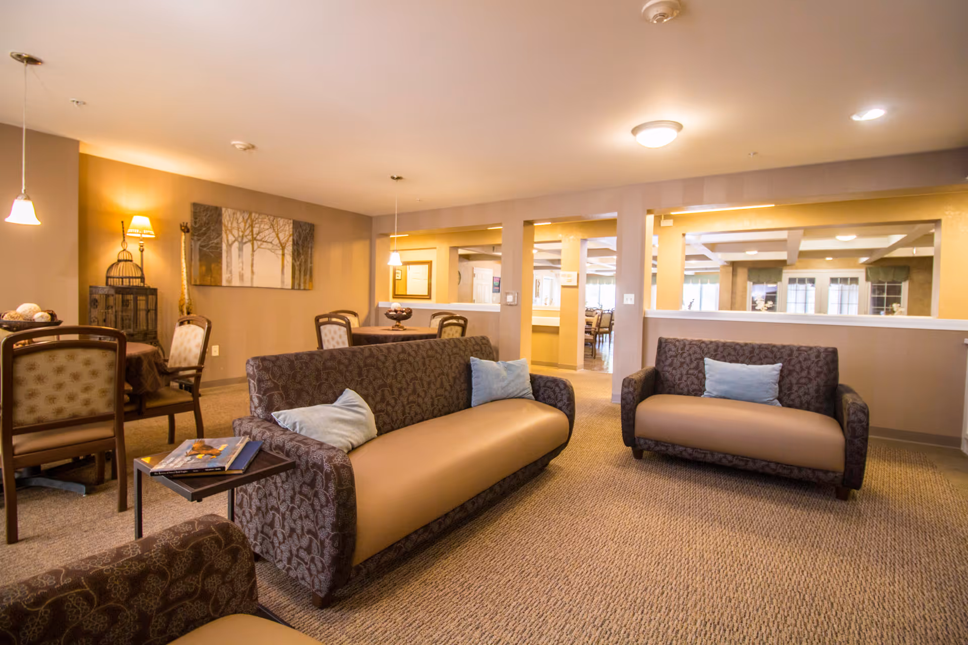 A cozy assisted living common area with two patterned sofas featuring beige cushions and light blue pillows, a small side table with magazines, and several dining tables with chairs in the background. The room is warmly lit with ceiling lights and wall lamps, and decorated with a nature-themed painting and a birdcage on a stand.