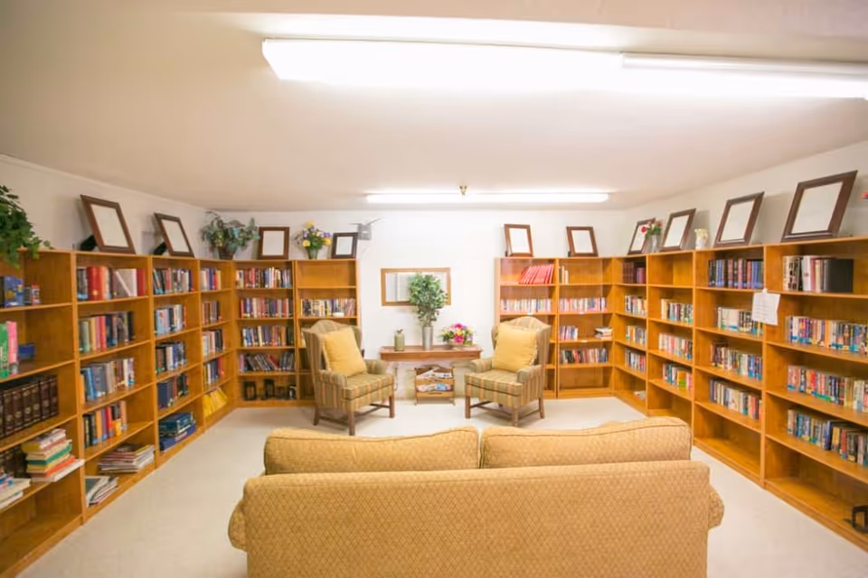 A cozy library room with wooden bookshelves lining the walls filled with books. Two cushioned armchairs with yellow pillows face a small wooden table with flowers and a plant. A beige sofa is positioned in the foreground, and the room is brightly lit with ceiling lights.