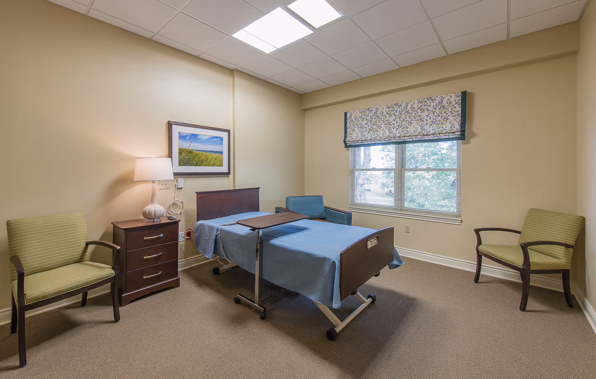 A simple room with a hospital-style bed covered with a blue blanket, a wooden bedside table with a lamp, two green armchairs, a blue armchair, a window with floral valance, and a framed picture of a beach scene on the wall.
