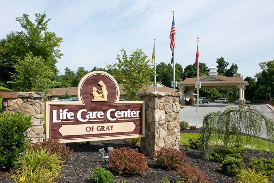Outdoor view of the entrance sign for Life Care Center of Gray, surrounded by landscaped greenery and stone pillars, with flags and a covered driveway in the background.