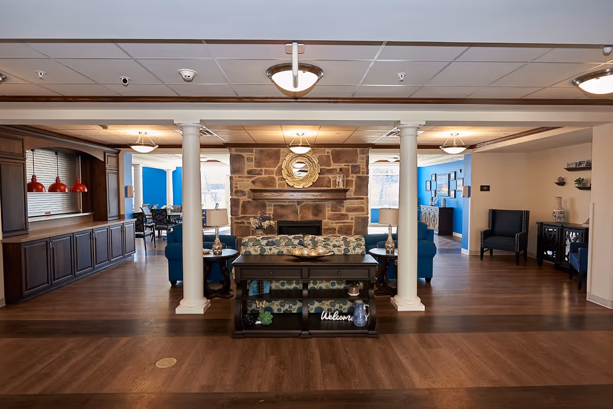 Spacious assisted living common area with seating, a stone fireplace, columns, and wood flooring.