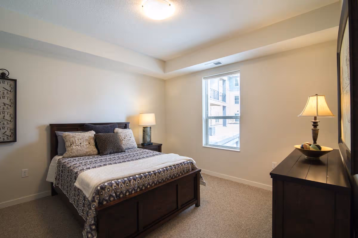 A cozy bedroom with a wooden bed frame, a patterned bedspread, and several pillows. There is a nightstand with a lamp on the left side of the bed and a dresser with a lamp and decorative bowl on the right side. A window lets in natural light, and a large clock hangs on the wall to the left.