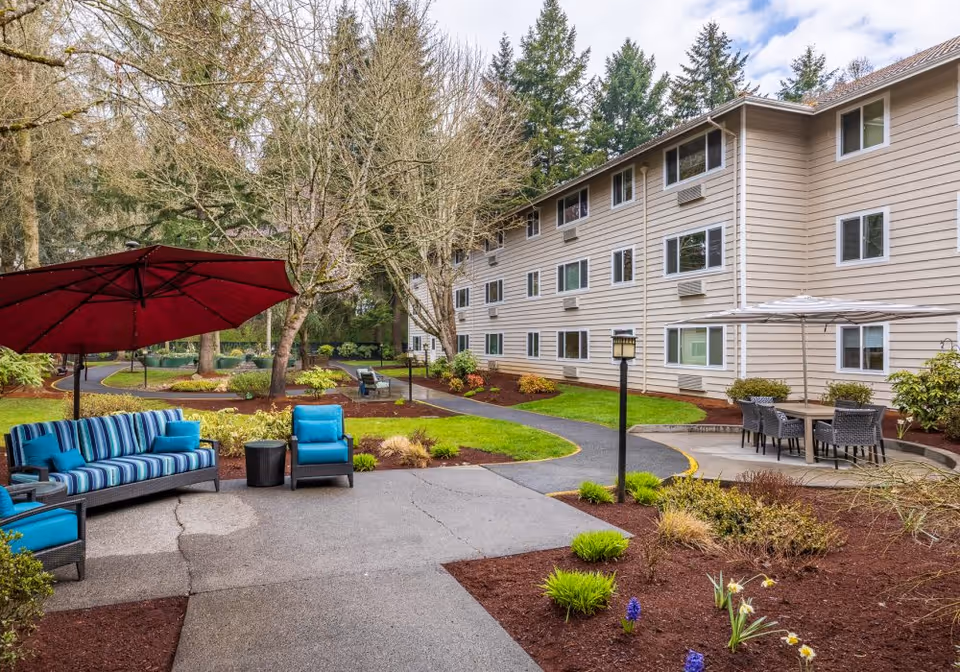 Outdoor patio area at Van Mall Retirement Vancouver featuring blue cushioned seating with a large red umbrella, a separate dining table with chairs under a white umbrella, surrounded by landscaped garden beds and a three-story beige building in the background.