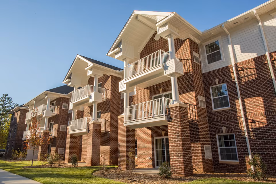 Brick three-story apartment building with balconies and a landscaped lawn under a clear blue sky.