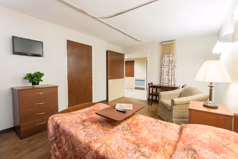 A cozy bedroom in a senior living facility featuring a bed with a patterned quilt, a wooden dresser with a small green plant, a wall-mounted TV, a cushioned armchair, a wooden side table with a lamp, and a small dining table with two chairs near a window with patterned curtains.