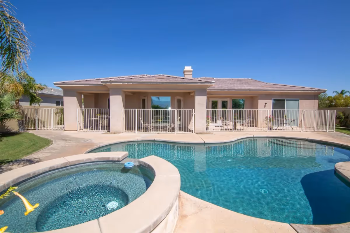 Single-story beige building with a fenced patio and a swimming pool with attached spa under a clear blue sky.