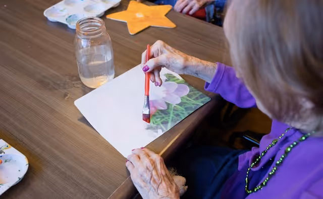 An elderly person wearing a purple sweater and green beaded necklace is painting a picture of a flower on a piece of paper at a wooden table. A jar of water and a star-shaped craft are also on the table.