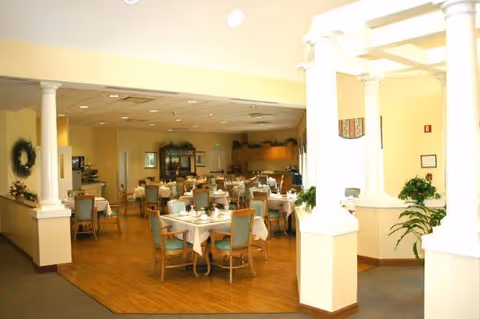 A spacious dining room with multiple tables covered with white tablecloths and set with dining ware. The room has wooden flooring, light yellow walls, and several chairs with light blue cushions. There are decorative plants and framed pictures on the walls, with white columns separating the dining area from the adjacent space.
