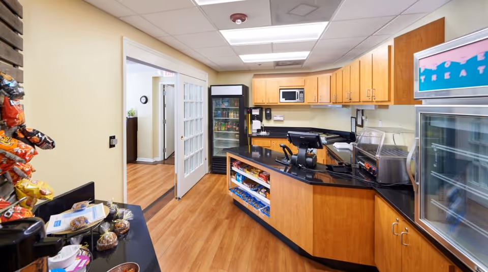 Interior view of a small snack and beverage area in a senior living facility named Galleria Woods, featuring wooden cabinets, a black countertop with a cash register, a glass-front refrigerator stocked with drinks, a microwave, and shelves stocked with snacks. The floor is wood laminate and there is a doorway with glass-paneled double doors leading to another room.