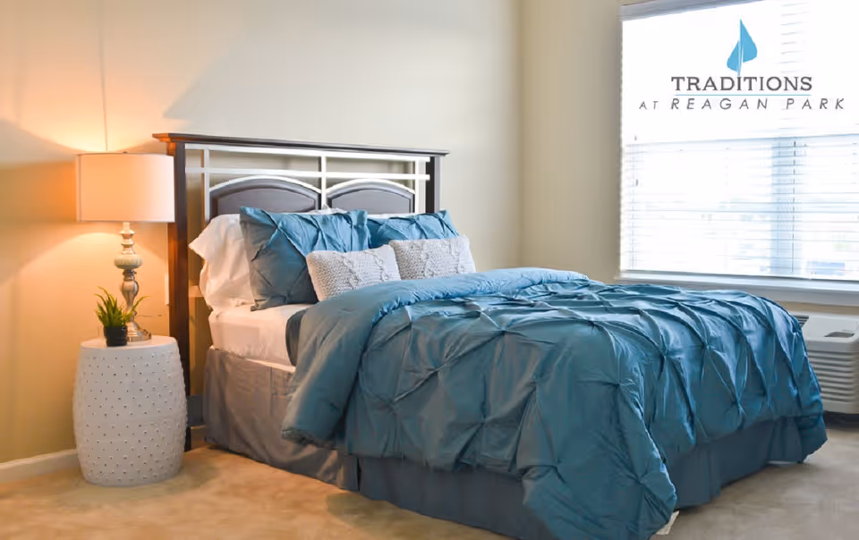 A neatly made bed with blue bedding and multiple pillows in a bedroom. There is a white nightstand with a small plant and a lamp on it next to the bed. A window with blinds is visible on the right side, and the room has beige walls and carpet. The logo for Traditions at Reagan Park is visible in the upper right corner.