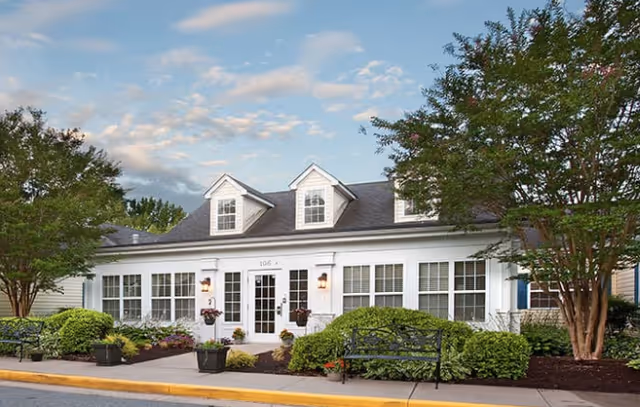 Exterior view of a single-story white building with large windows and a central entrance door, surrounded by well-maintained bushes, trees, and flower pots under a partly cloudy sky.