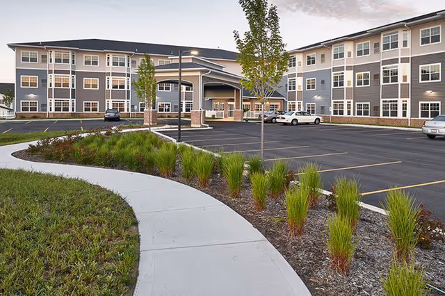 Exterior view of a modern senior living facility with a curved sidewalk, landscaped greenery, and a parking lot with several cars parked near the building under a partly cloudy sky.